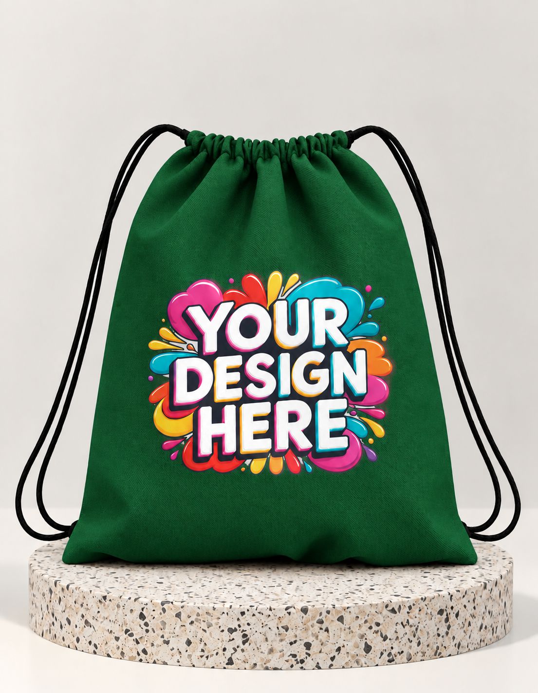 A green drawstring bag with black cords, featuring a colorful 'YOUR DESIGN HERE' graphic on the front, displayed on a round speckled stone riser against a light gray background.