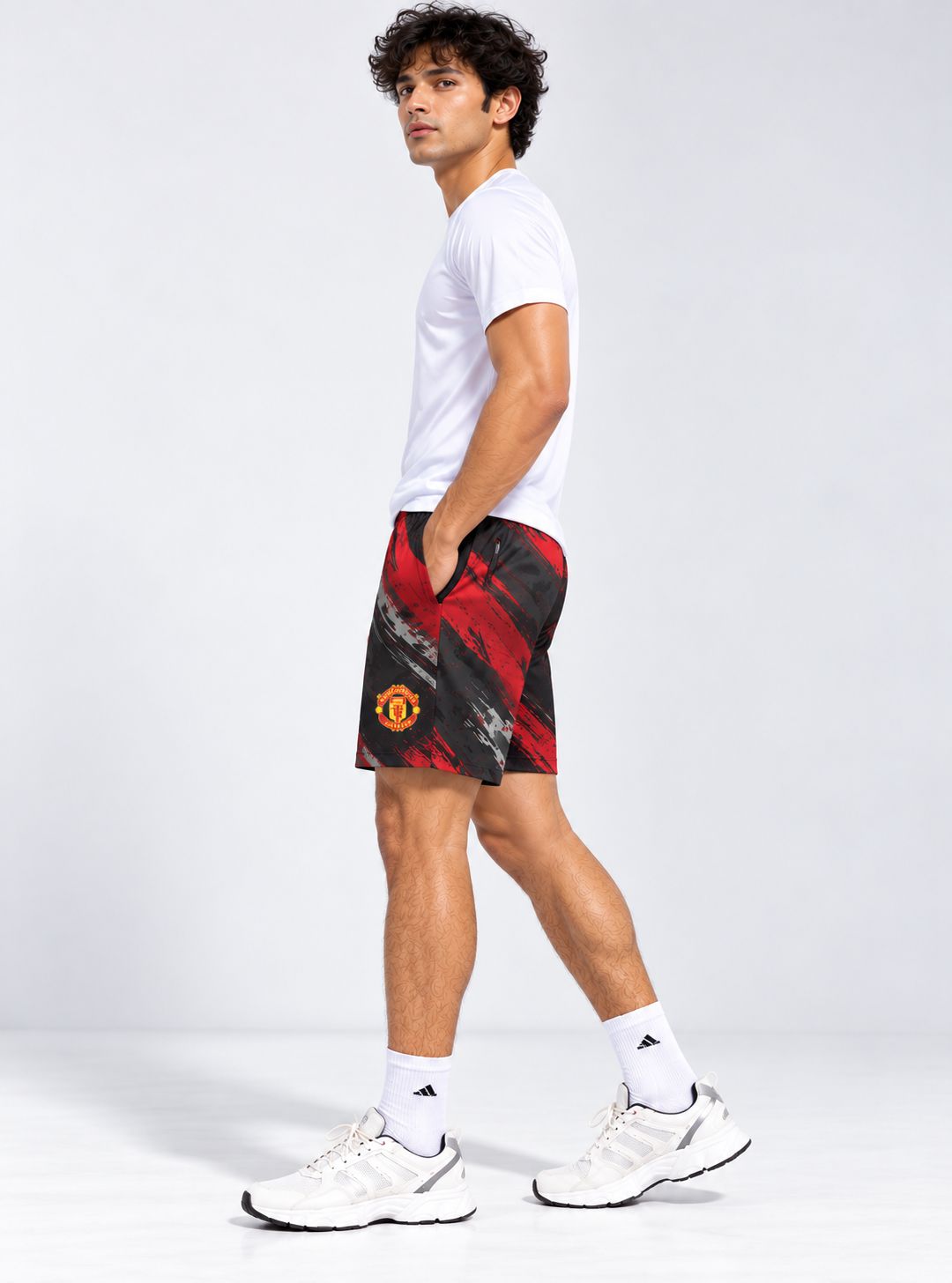 A man in a white t-shirt and white sneakers models a pair of red and black Manchester United athletic shorts, standing in a side profile against a light gray studio background.