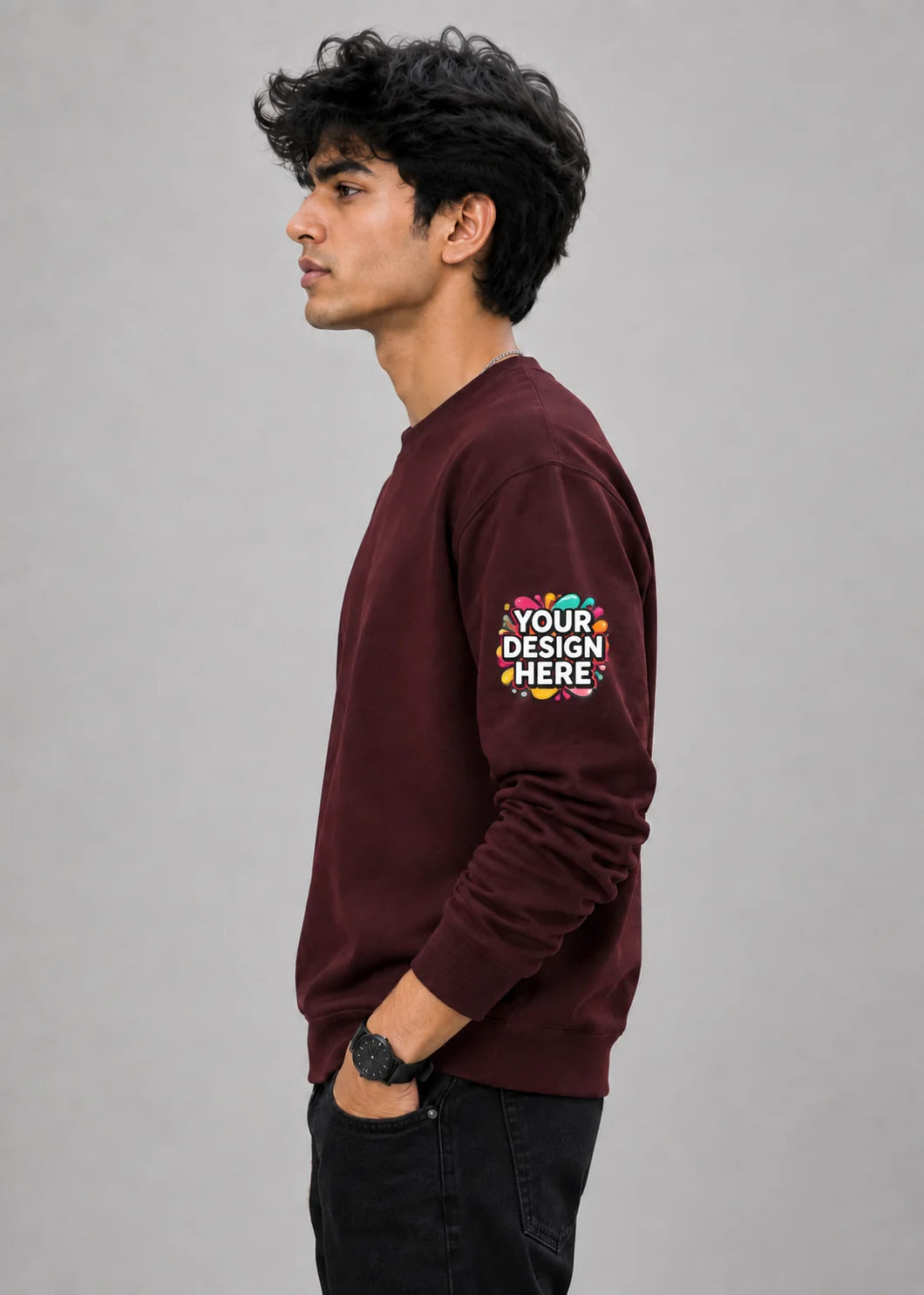A man in profile wearing a maroon crewneck sweatshirt with a colorful 'YOUR DESIGN HERE' mockup graphic on the left sleeve, paired with black pants, against a solid gray studio background.