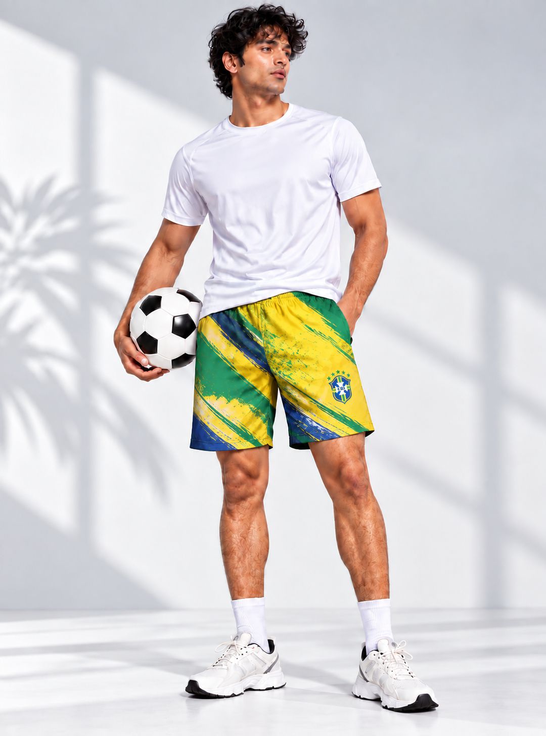 A man modeling athletic shorts with a vibrant green, yellow, and blue abstract pattern, styled with a white t-shirt, white socks, and white sneakers, while holding a black and white soccer ball against a white wall with soft shadows.