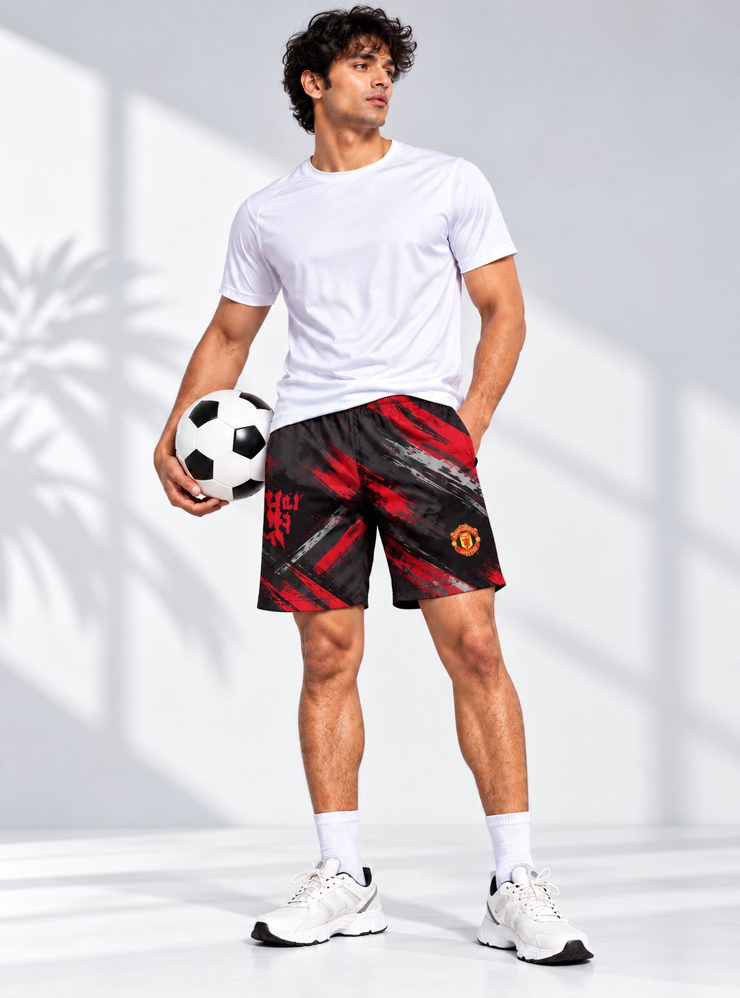 A man modeling black and red Manchester United athletic shorts, paired with a white t-shirt, white socks, and white sneakers, holding a soccer ball against a bright studio background with soft shadows.