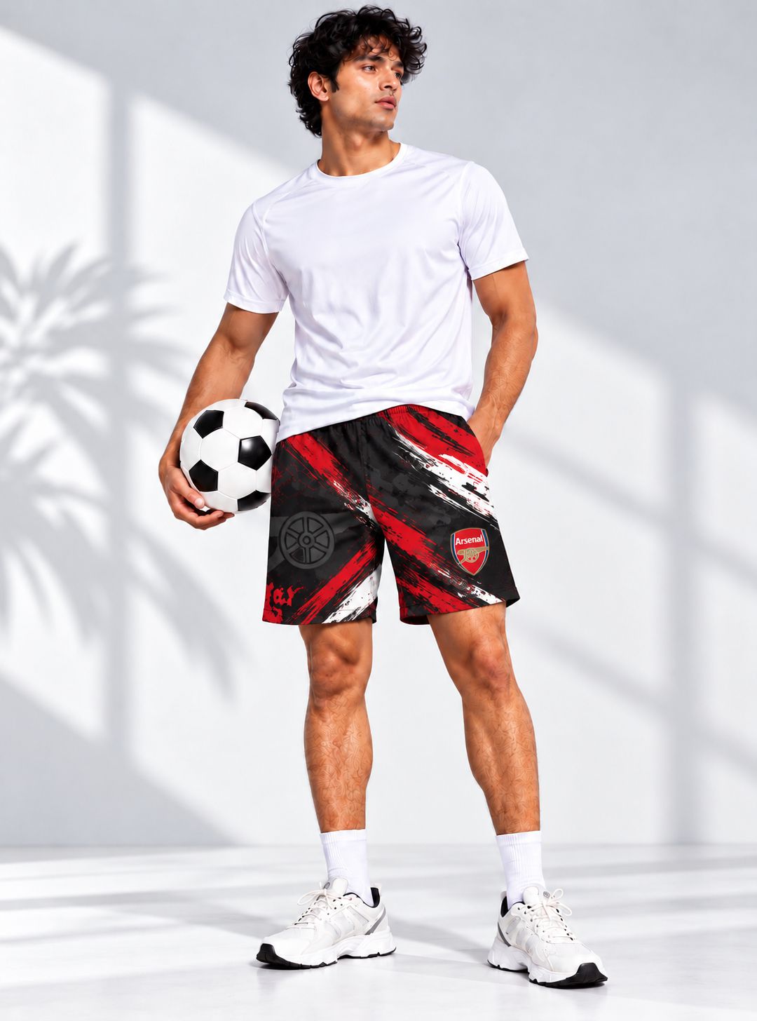 A man modeling black and red patterned soccer shorts with Arsenal branding, paired with a white t-shirt, white socks, and white sneakers, holding a soccer ball against a bright studio background with soft shadow patterns.