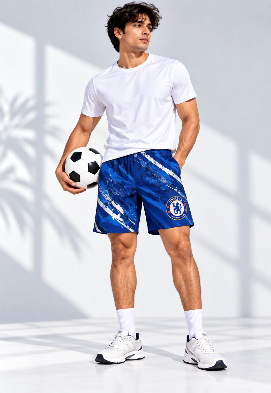 A man modeling blue and white Chelsea FC athletic shorts, paired with a white t-shirt, white socks, and white sneakers, holding a soccer ball against a bright studio background with soft shadows.