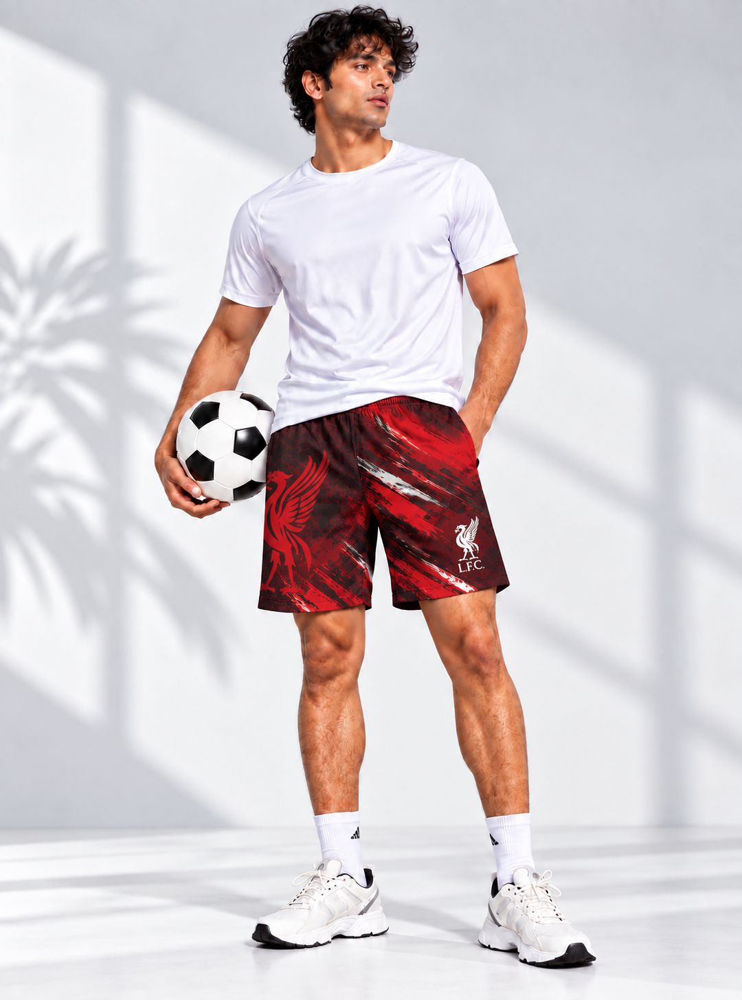 A man modeling red and black patterned athletic shorts with a white logo, paired with a white t-shirt, white socks, and white sneakers, holding a soccer ball in a studio setting with shadow patterns.
