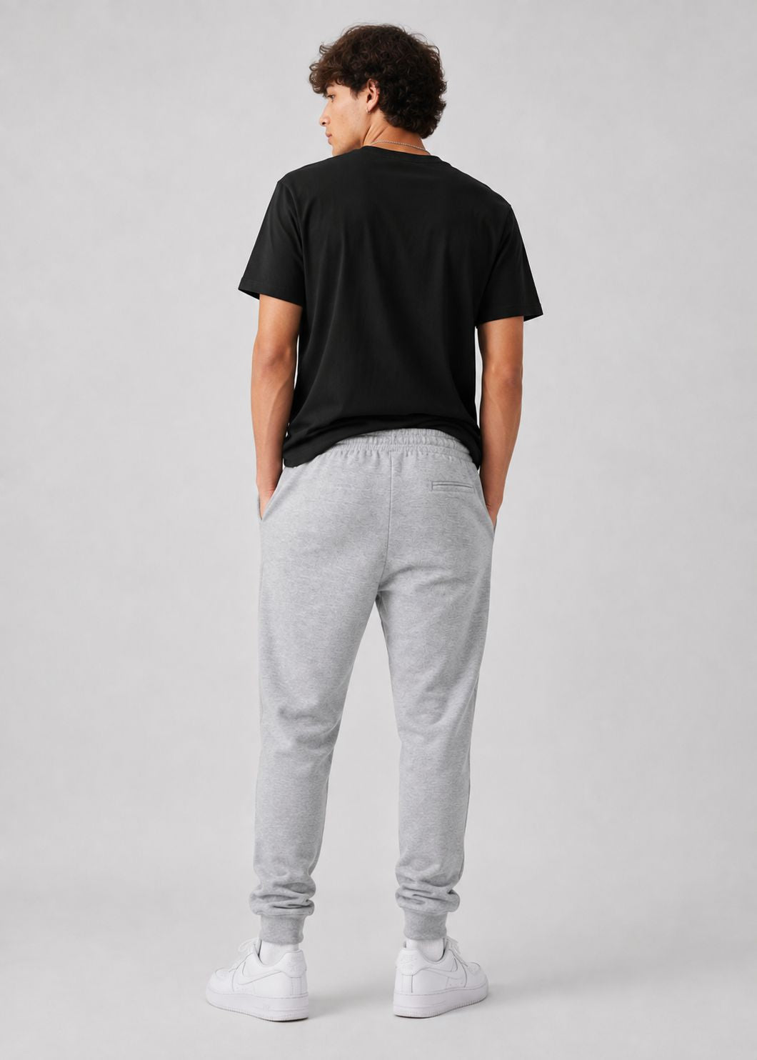 A man shown from behind wearing a black t-shirt, light gray heather sweatpants, and white sneakers against a light gray studio background.