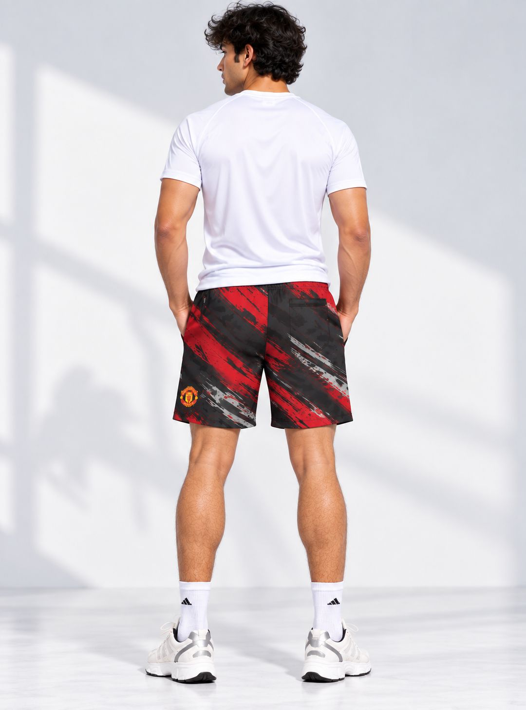 A man viewed from behind wearing Manchester United athletic shorts with a red and black abstract pattern, a white t-shirt, white socks, and white sneakers in a bright studio setting.