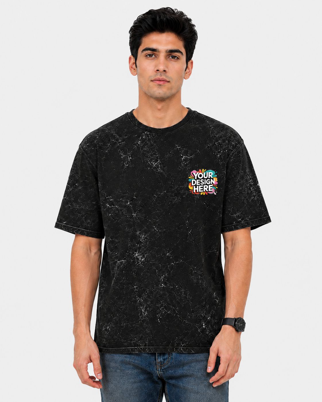 A man wearing a black mineral-wash t-shirt with a colorful 'YOUR DESIGN HERE' placeholder graphic on the left chest, paired with blue jeans, against a light gray background.