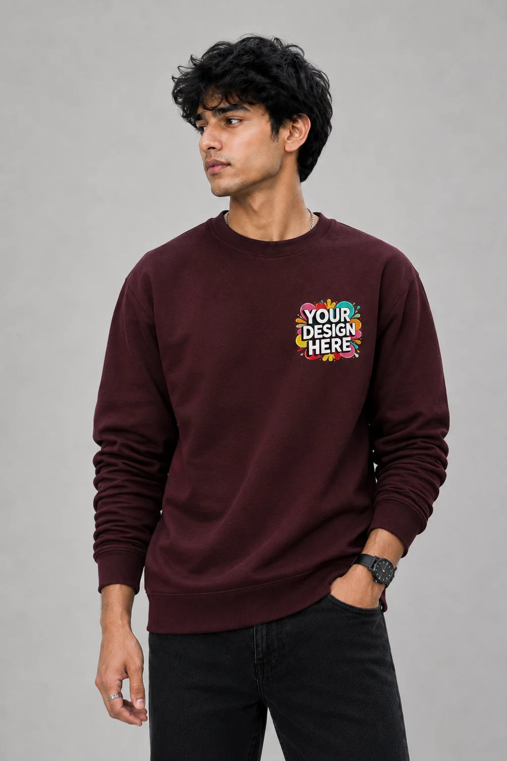 A man wearing a maroon crewneck sweatshirt with a colorful 'YOUR DESIGN HERE' mockup graphic on the left chest, paired with black jeans, against a solid gray studio background.