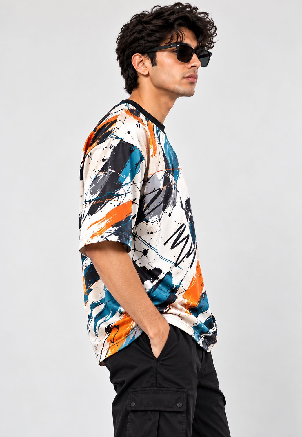 A man wearing a short-sleeve crewneck t-shirt with an abstract multicolor paint splatter pattern in orange, blue, black, and white, paired with black cargo pants and sunglasses, set against a plain light gray background.