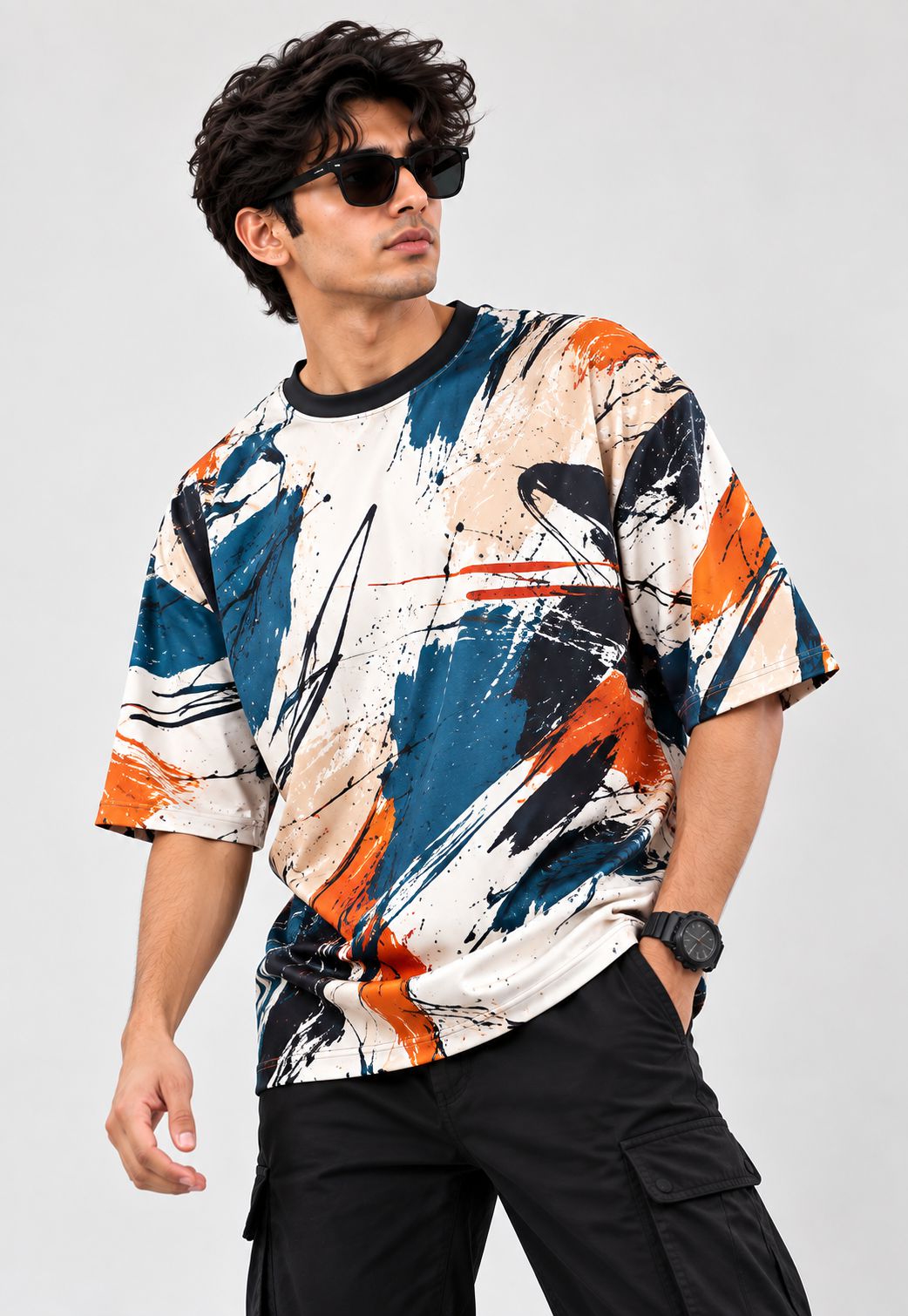 A man wearing a short-sleeve crewneck t-shirt with an abstract paint-splatter pattern in blue, orange, black, and beige, paired with black cargo pants and sunglasses, against a light gray studio background.