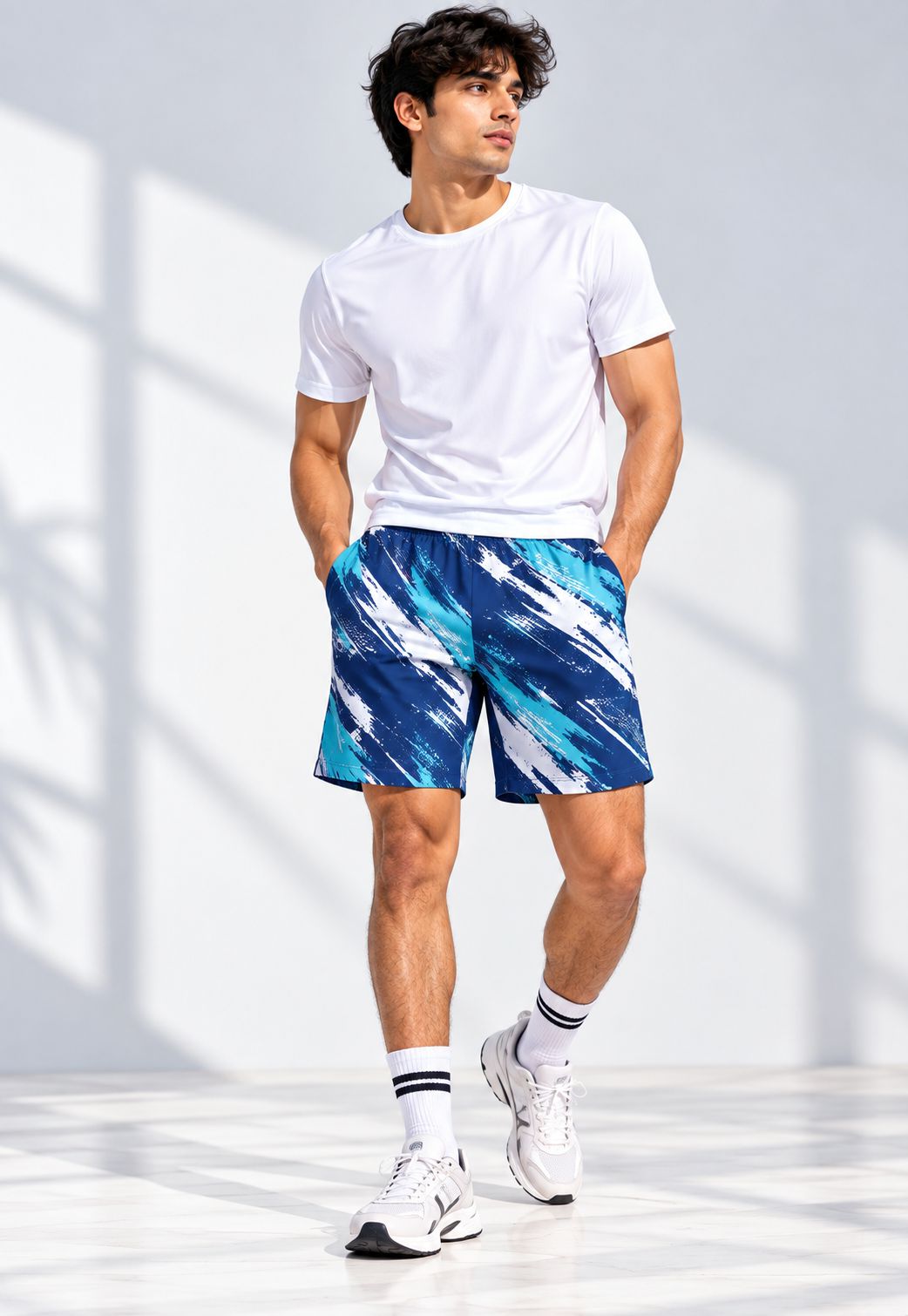 A man wearing a white t-shirt, blue and white patterned athletic shorts, white socks, and white sneakers, standing in a bright studio with window shadow patterns on a white wall.