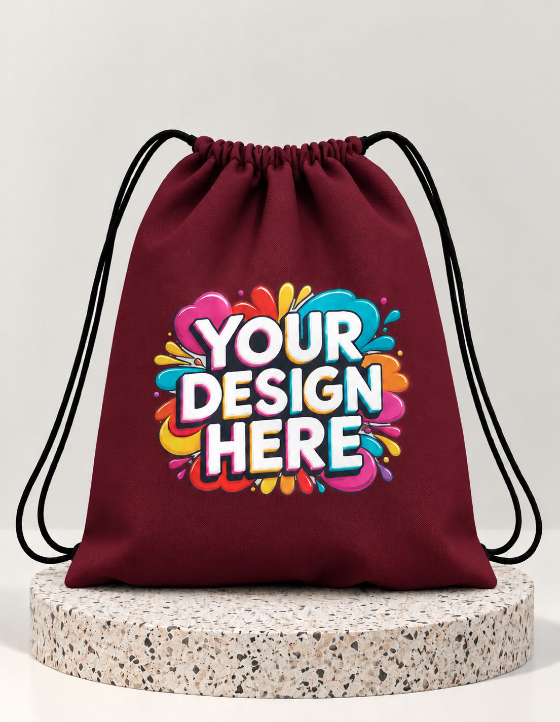 A maroon drawstring bag with black cords, featuring a colorful 'YOUR DESIGN HERE' mockup graphic on the front, displayed on a round speckled stone pedestal against a light gray background.
