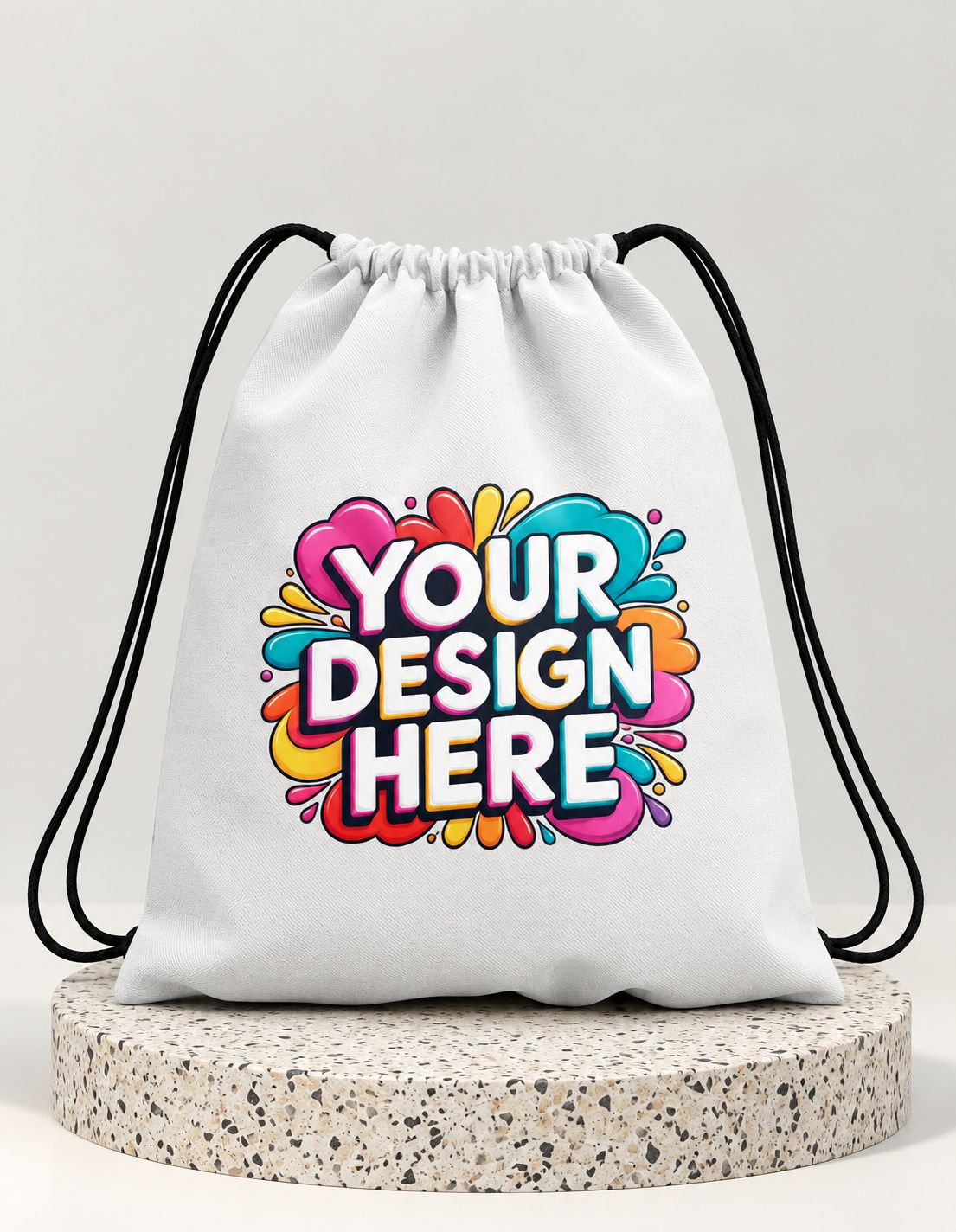 A white drawstring bag with black cords, featuring a colorful 'YOUR DESIGN HERE' graphic on the front, displayed on a round speckled pedestal against a light gray background.