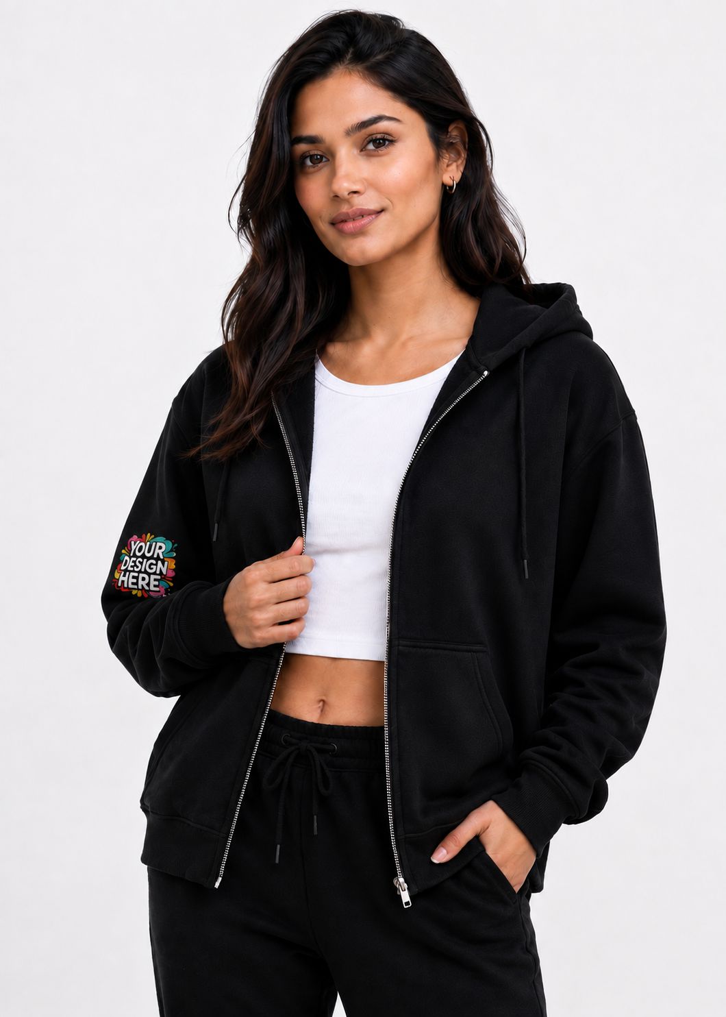 A woman modeling a black zip-up hoodie with a colorful 'YOUR DESIGN HERE!' graphic on the left sleeve, worn over a white crop top and black sweatpants against a light gray background.