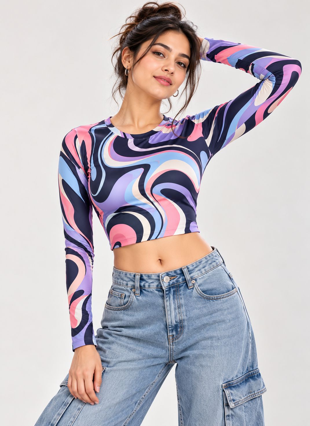 A woman modeling a long-sleeve crop top with a colorful abstract swirl pattern in pink, purple, blue, and black, paired with light-wash blue jeans against a light gray studio background.