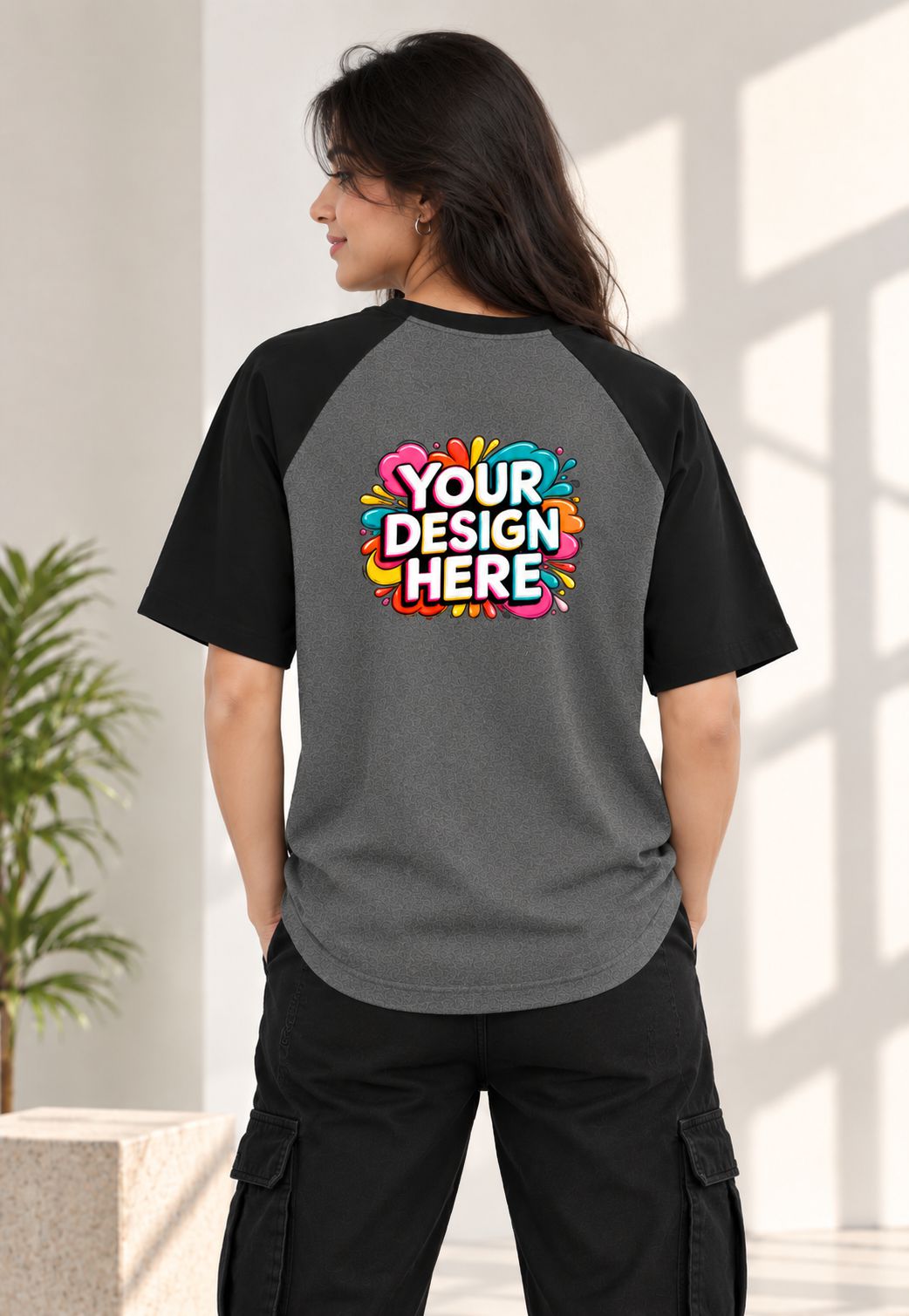 A woman shown from the back wearing a custom raglan-style t-shirt. The shirt has a heather gray body and black sleeves, featuring a colorful 'YOUR DESIGN HERE' mockup graphic on the back.