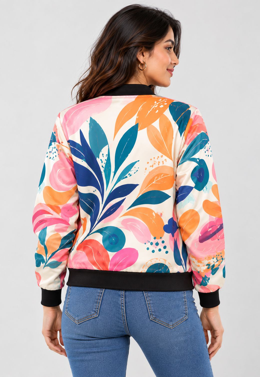 A woman shown from the back wearing a long-sleeve bomber jacket with a colorful abstract floral pattern in pink, orange, teal, and blue on a cream background, paired with blue jeans against a light gray studio background.