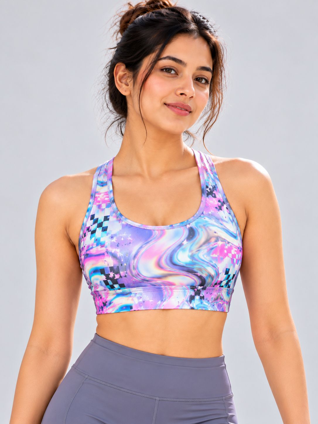 A woman wearing a colorful sports bra with a purple, blue, and pink abstract pattern, paired with gray athletic leggings, against a solid light gray background.