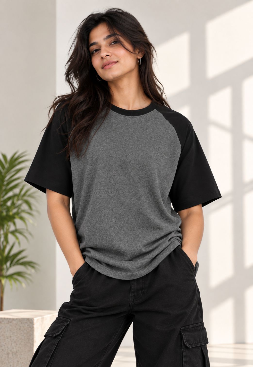 A woman wearing a heather gray and black raglan t-shirt with black cargo pants, standing in a brightly lit room with window shadows.