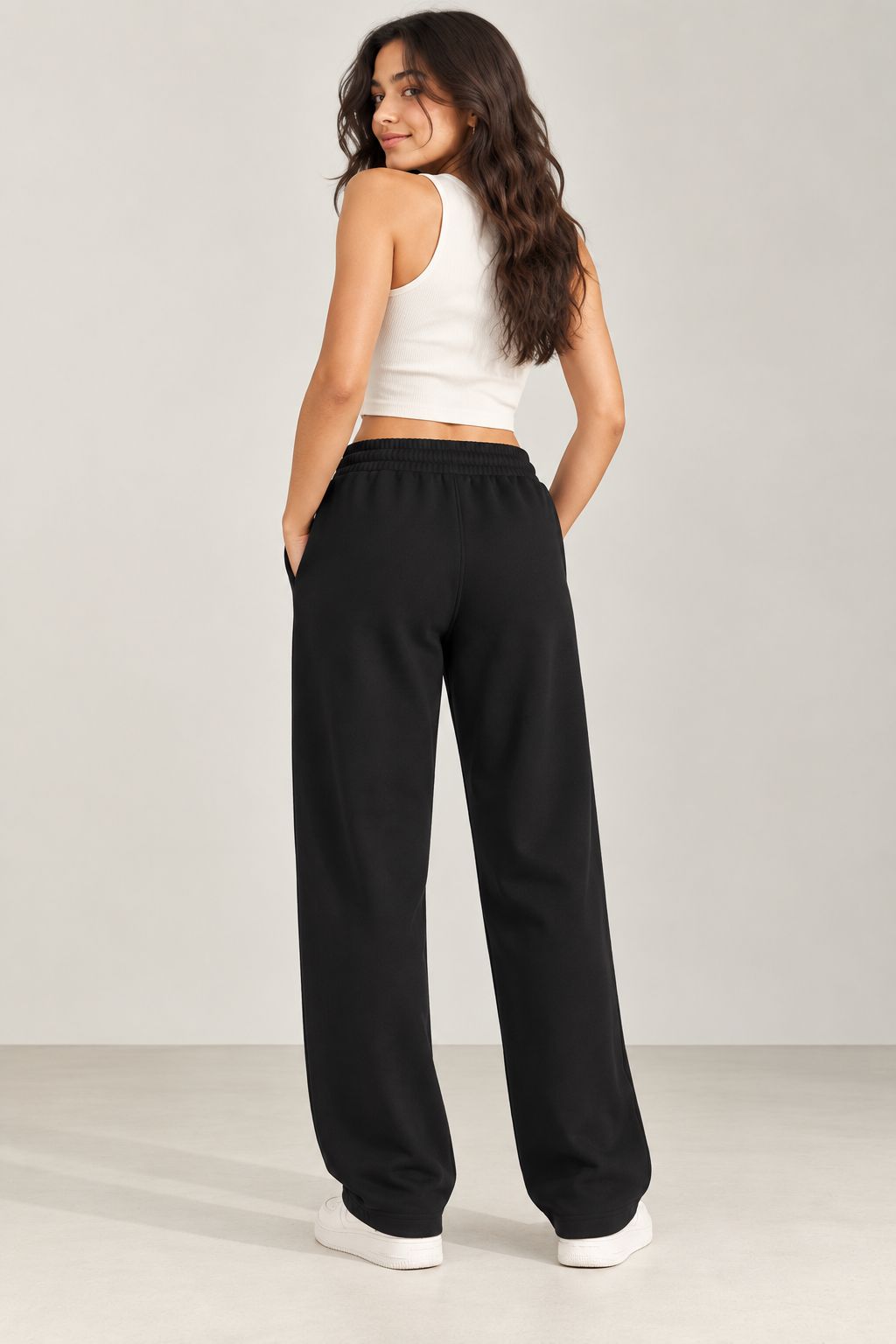 A woman wearing black wide-leg sweatpants, a white ribbed tank top, and white sneakers, photographed from the back against a light gray studio background.