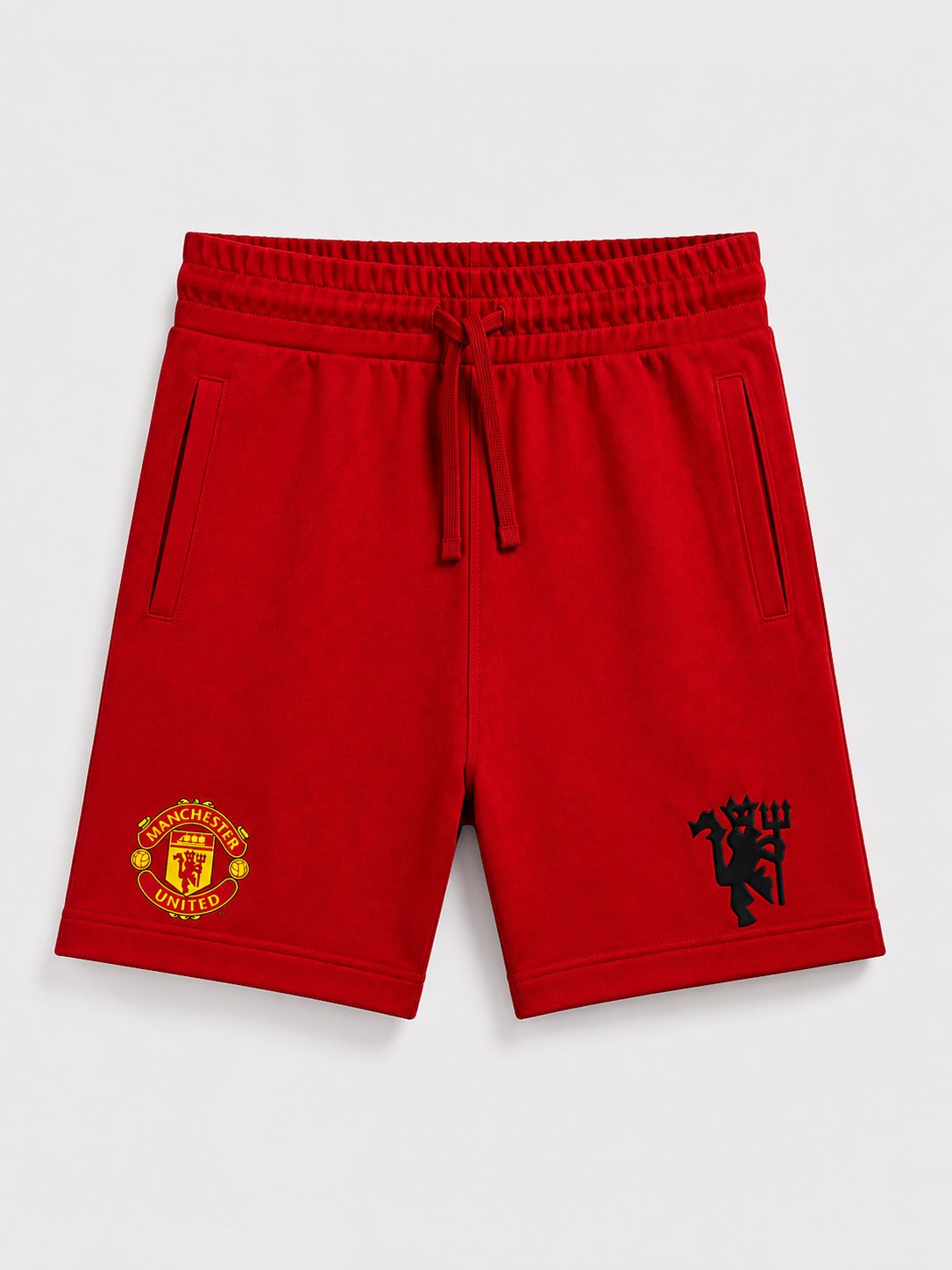 Red Manchester United athletic shorts with a drawstring waist, side pockets, and embroidered club crests on the left and right legs, shown on a light gray background.