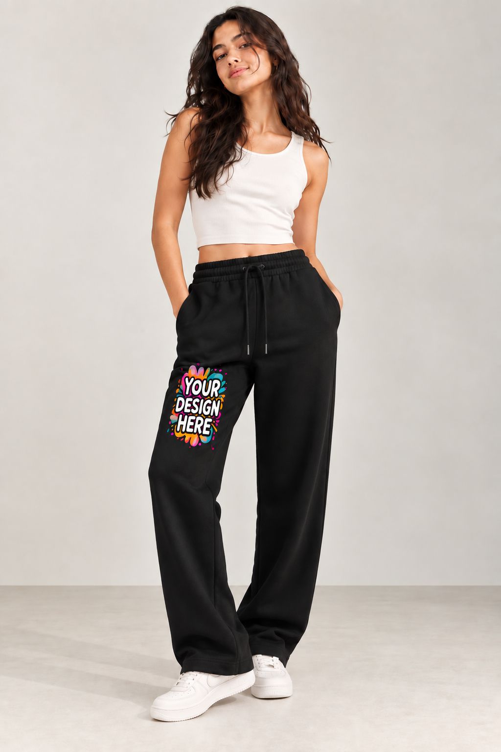 A woman modeling black wide-leg sweatpants with a colorful 'YOUR DESIGN HERE' mockup graphic on the left thigh, paired with a white crop top and white sneakers, against a light gray studio background.