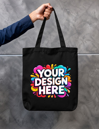 A black canvas tote bag with long handles, held by a person's hand against a gray textured wall. The bag features a colorful graphic with the text 'YOUR DESIGN HERE' in white with a black outline, surrounded by a vibrant multicolor paint splatter design.