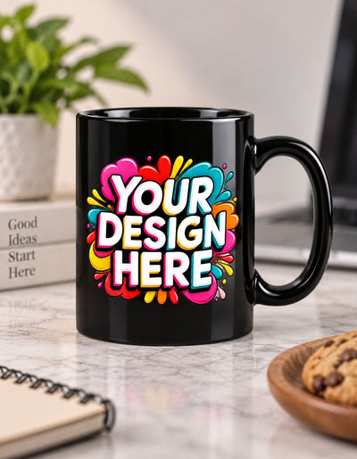A black ceramic mug with a colorful 'YOUR DESIGN HERE' graphic on the front, set on a marble countertop with a blurred background showing a plant, notebook, and cookie.
