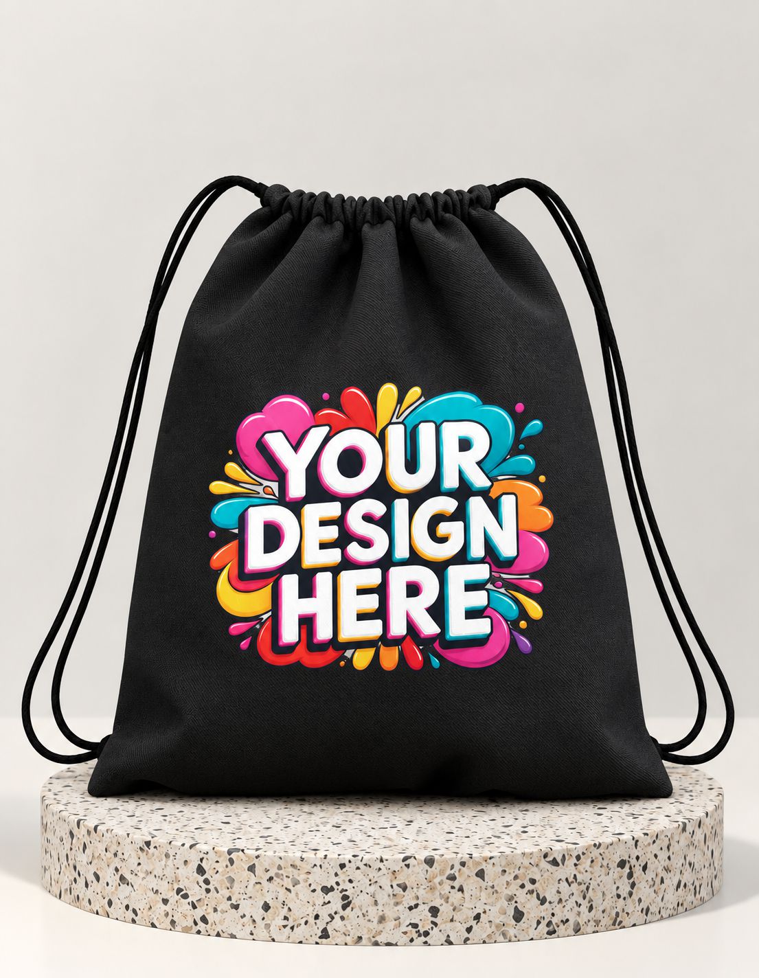 A black drawstring bag with black cords, featuring a colorful graphic print that reads 'YOUR DESIGN HERE' in white letters with a pink, blue, yellow, and orange paint splatter background. The bag is displayed on a round speckled stone pedestal against a light gray background.
