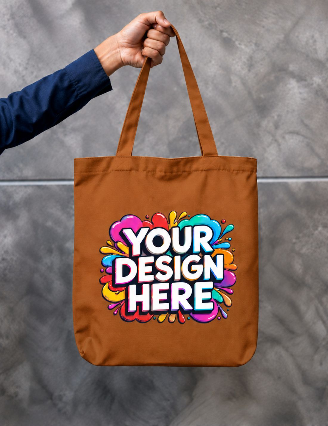 A brown canvas tote bag held by a person's hand against a gray concrete wall. The bag features a colorful abstract paint splatter graphic with the text 'YOUR DESIGN HERE' in white with a black outline.