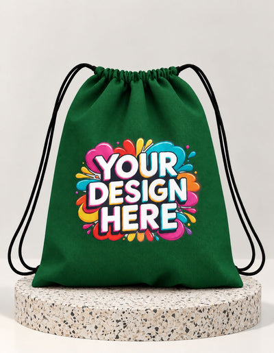 A green drawstring bag with black cords, featuring a colorful 'YOUR DESIGN HERE' graphic on the front, displayed on a round speckled stone riser against a light gray background.