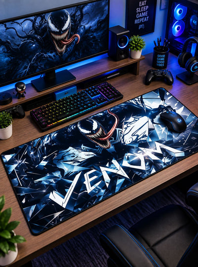 A large desk mat featuring Venom artwork in blue, black, and white, styled on a wooden desk in a gaming setup with a keyboard, mouse, monitor, and RGB lighting.