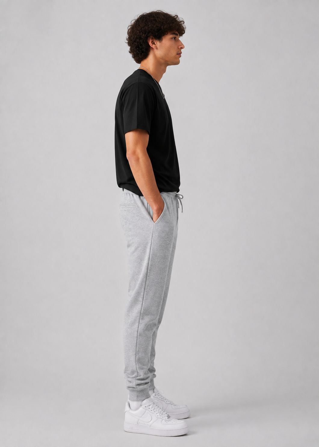 A man in profile wearing a black t-shirt, light gray jogger sweatpants, and white sneakers, standing against a neutral light gray studio background.