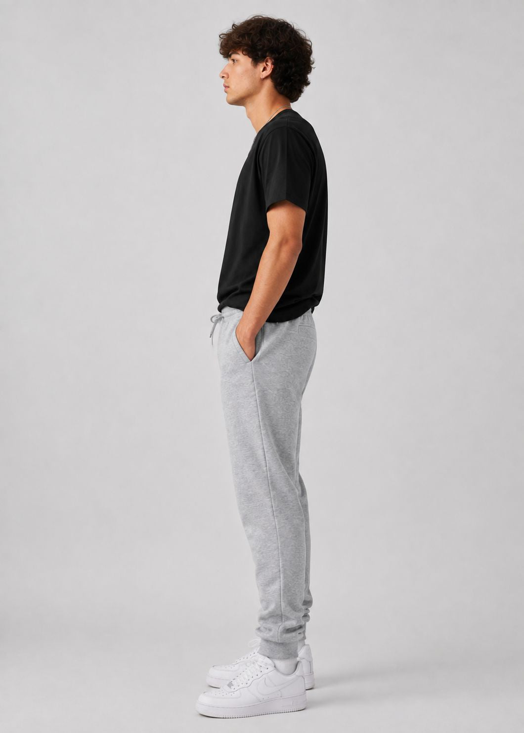 A man in profile wearing a black t-shirt, light gray sweatpants, and white sneakers, standing against a solid light gray studio background.