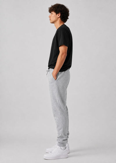 A man in profile wearing a black t-shirt, light gray sweatpants, and white sneakers, standing against a solid light gray studio background.