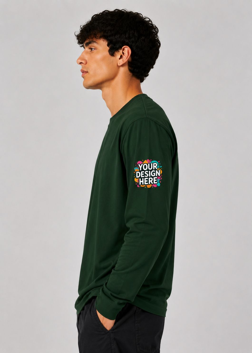 A man in profile wearing a dark green long-sleeve t-shirt with a colorful mockup graphic on the left sleeve that reads 'YOUR DESIGN HERE', set against a plain light gray studio background.