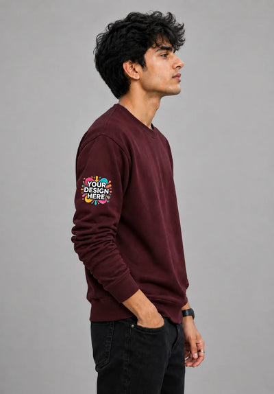 A man in profile wearing a maroon crewneck sweatshirt with a colorful 'YOUR DESIGN HERE' mockup graphic on the left sleeve, paired with black jeans, against a solid gray background.