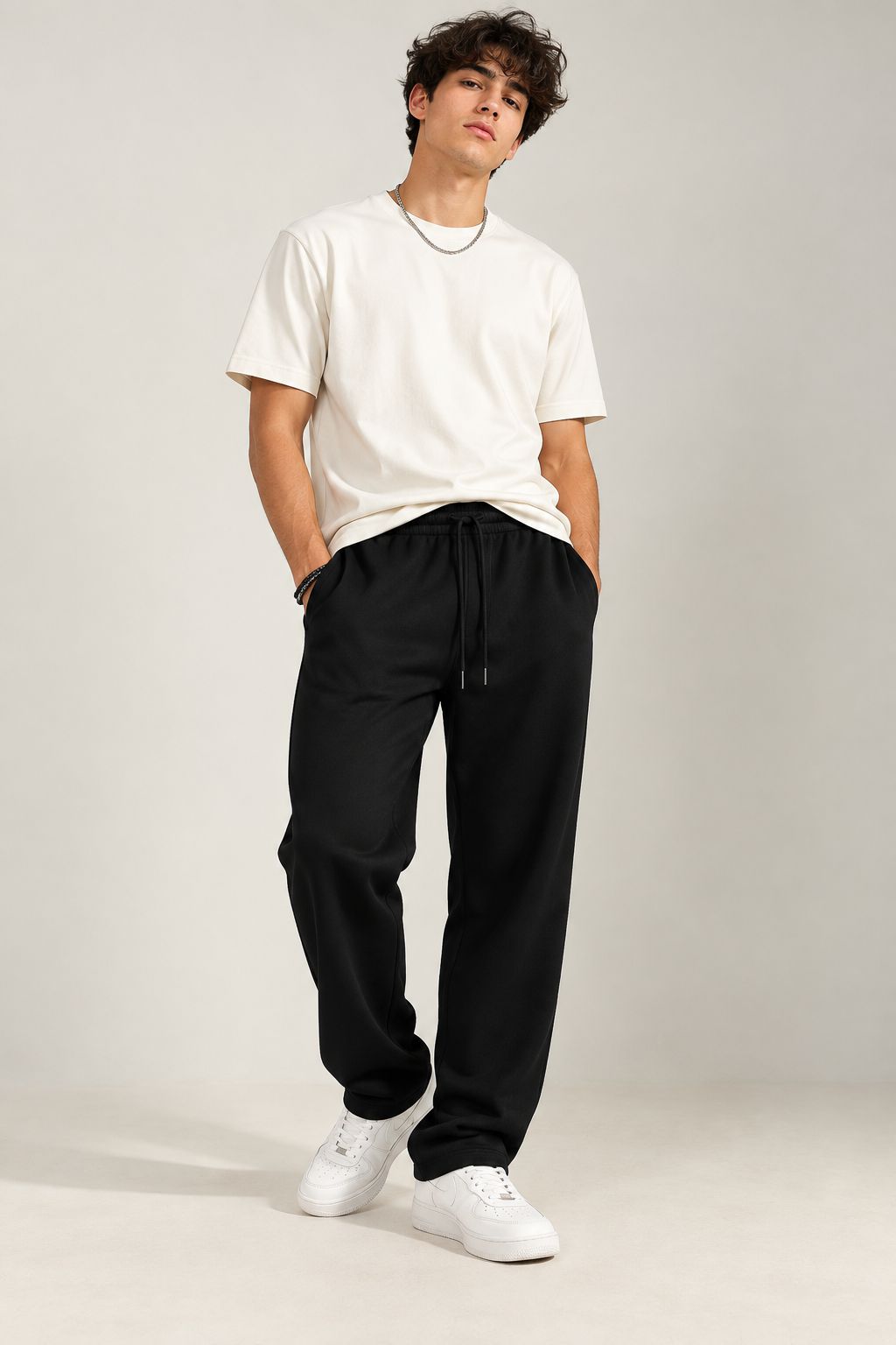 A man modeling a white crewneck t-shirt and black drawstring sweatpants, paired with white sneakers, against a neutral light gray studio background.