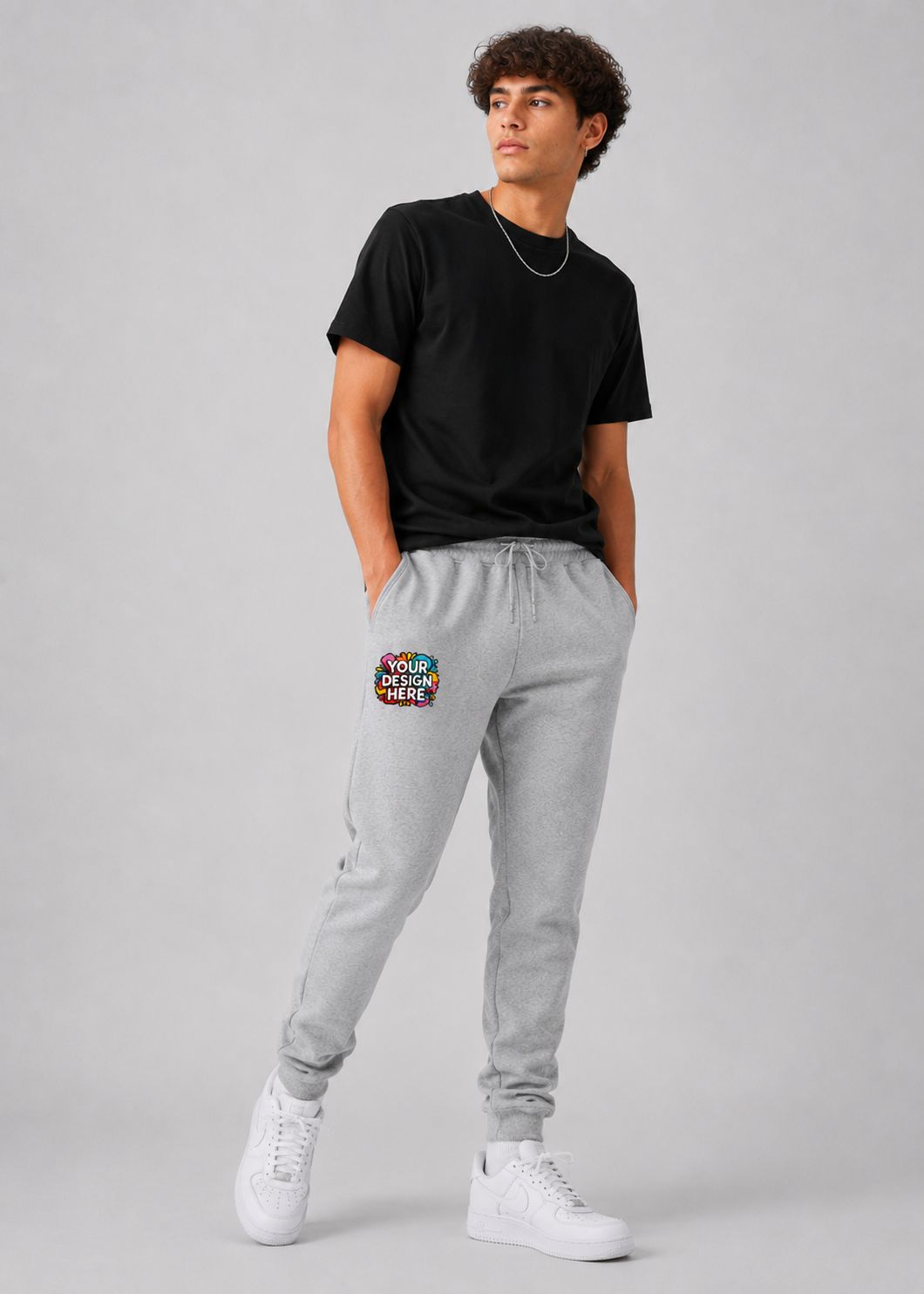 A man modeling light gray jogger sweatpants with a colorful graphic patch on the left thigh, paired with a black t-shirt and white sneakers, against a light gray studio background.