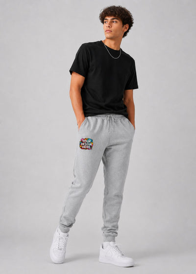 A man modeling light gray jogger sweatpants with a colorful graphic patch on the left thigh, paired with a black t-shirt and white sneakers, against a light gray studio background.