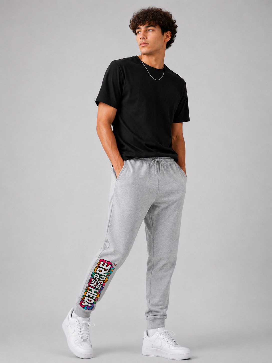 A man modeling light gray sweatpants with a colorful graphic print down the side, paired with a black t-shirt and white sneakers, against a solid gray studio background.