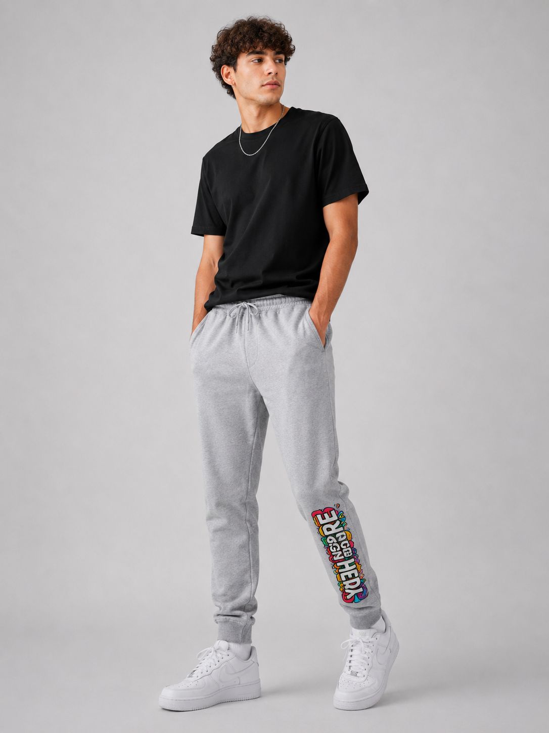 A man modeling light gray sweatpants with a colorful graphic print on the lower left leg, paired with a black t-shirt and white sneakers, against a solid gray studio background.