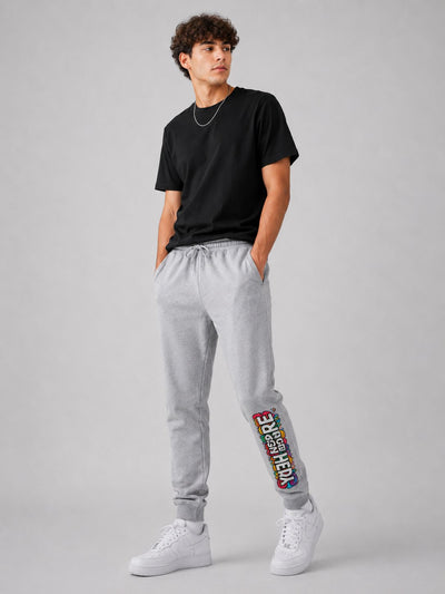 A man modeling light gray sweatpants with a colorful graphic print on the lower left leg, paired with a black t-shirt and white sneakers, against a solid gray studio background.