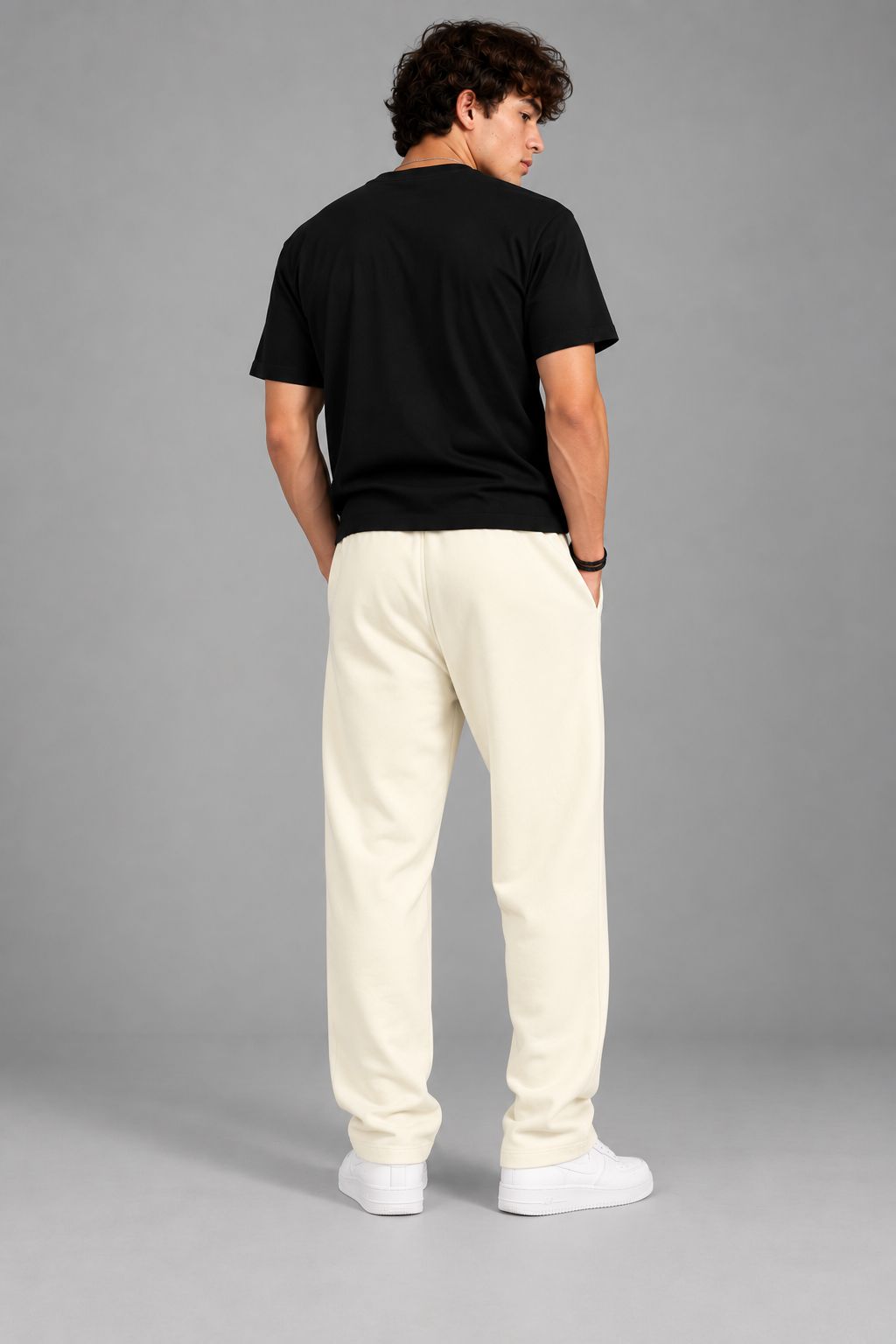 A man shown from behind wearing a black t-shirt, cream-colored sweatpants, and white sneakers, standing against a solid gray studio background.