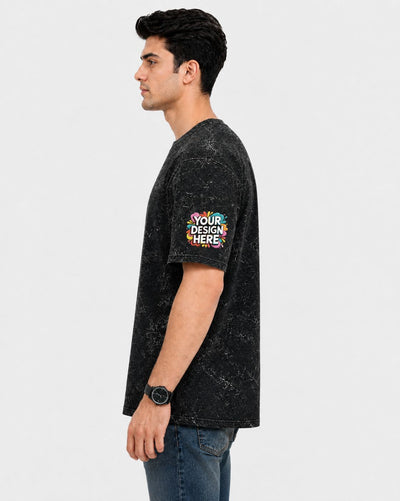 A man shown in profile wearing a black mineral-wash t-shirt with a colorful 'YOUR DESIGN HERE' mockup graphic on the left sleeve, paired with blue jeans against a light gray background.