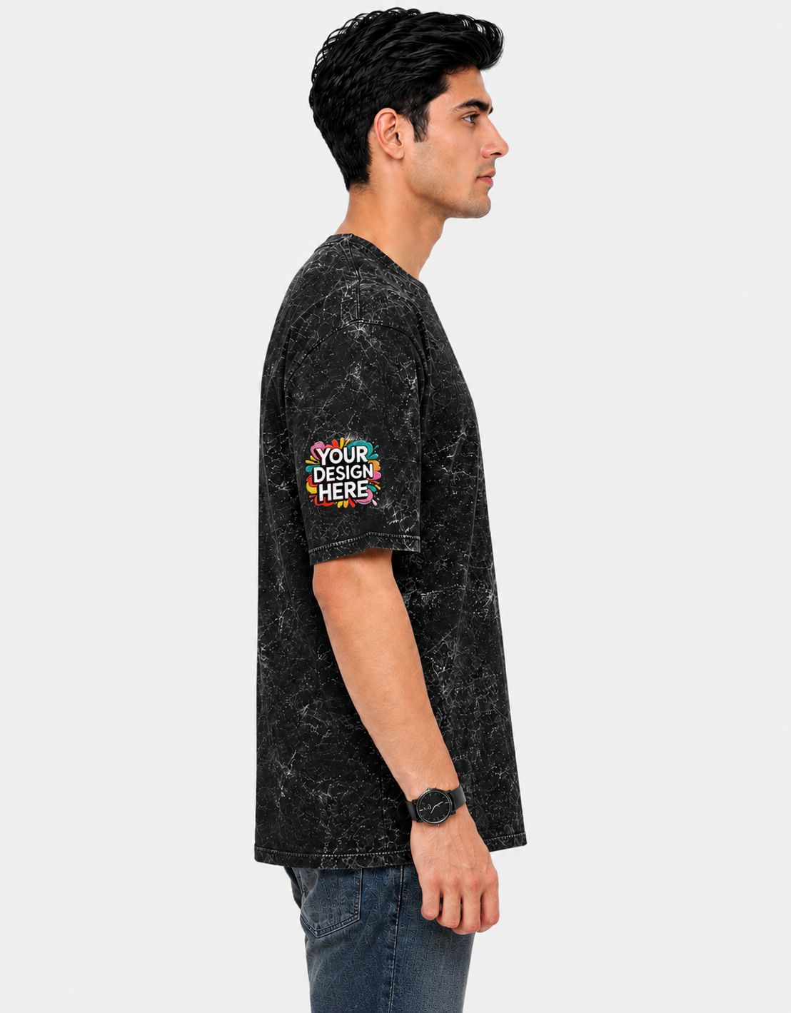 A man shown in profile wearing a black mineral-wash t-shirt with a colorful 'YOUR DESIGN HERE' mockup patch on the sleeve, paired with blue jeans against a light gray background.