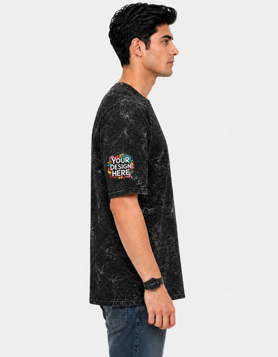 A man shown in profile wearing a black mineral-wash t-shirt with a colorful 'YOUR DESIGN HERE' mockup patch on the sleeve, paired with blue jeans against a light gray background.