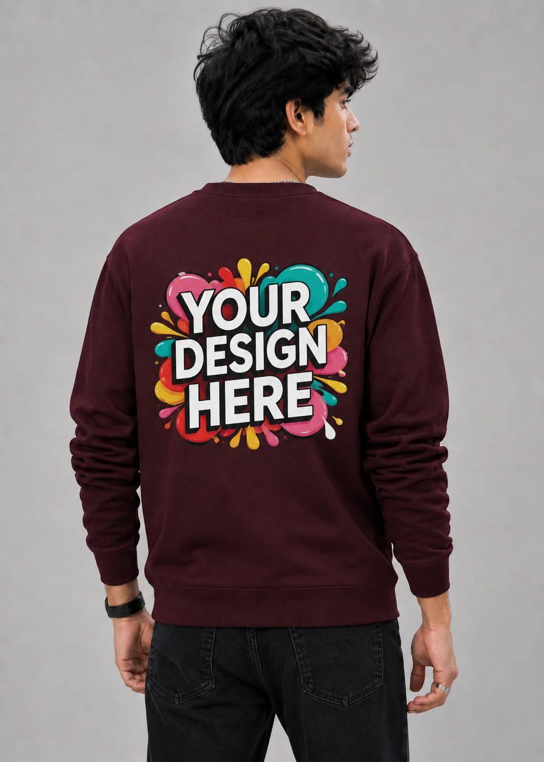 A man viewed from behind wearing a maroon crewneck sweatshirt with a colorful 'YOUR DESIGN HERE' mockup graphic on the back, paired with black jeans against a neutral gray background.
