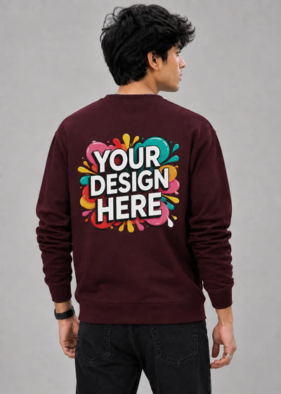 A man viewed from behind wearing a maroon crewneck sweatshirt with a colorful 'YOUR DESIGN HERE' mockup graphic on the back, paired with black jeans against a neutral gray background.