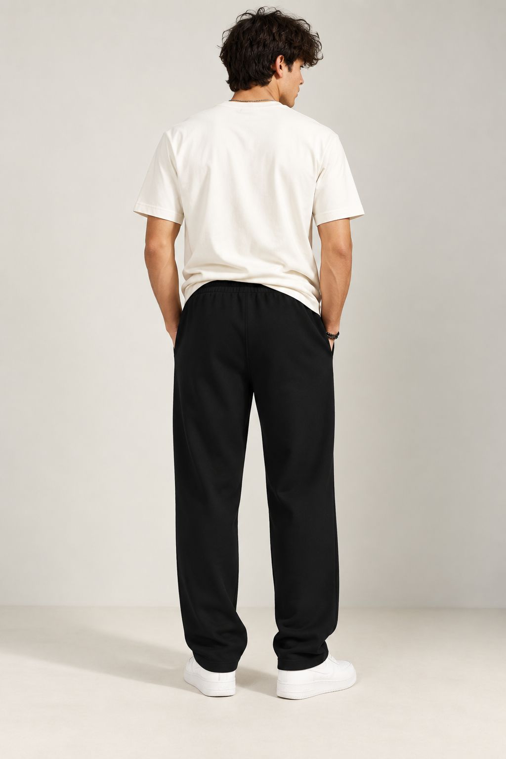 A man viewed from behind wearing a white t-shirt, black sweatpants, and white sneakers, standing against a neutral light gray studio background.